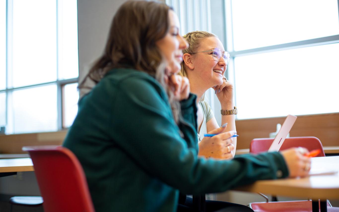Two students are sitting in a classroom, looking off into the distance and smiling